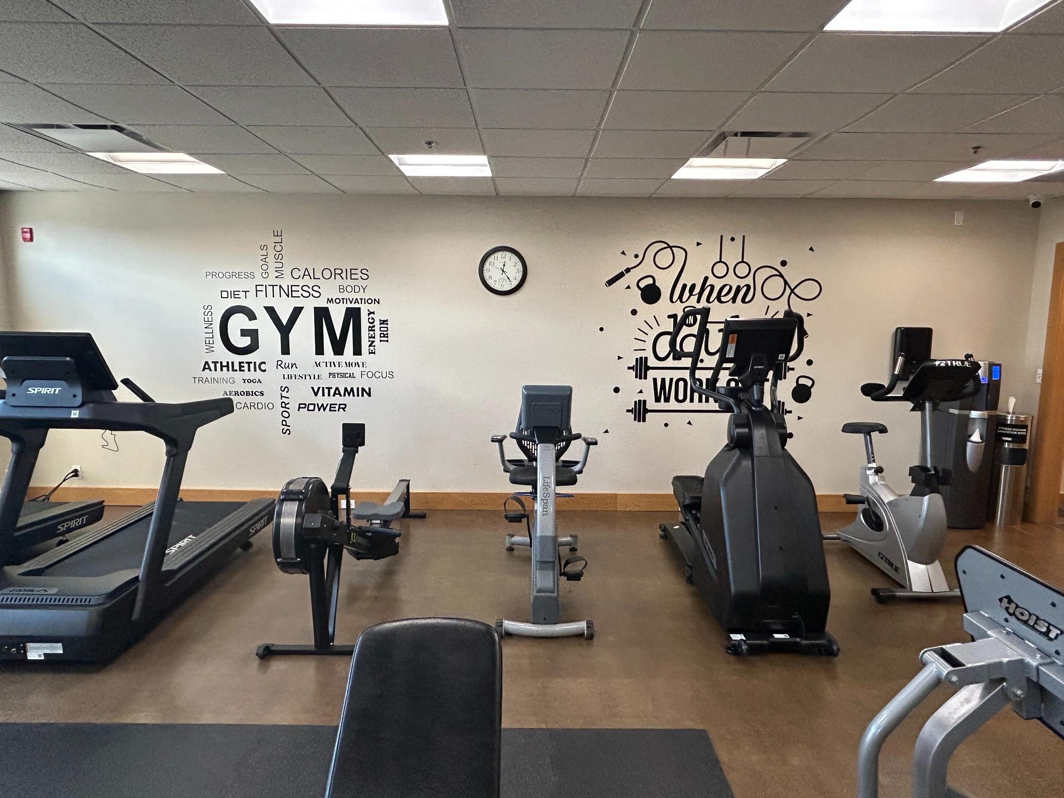 A small, well-equipped gym with cardio and strength machines against a white wall decorated with fitness-themed motivational text decals.