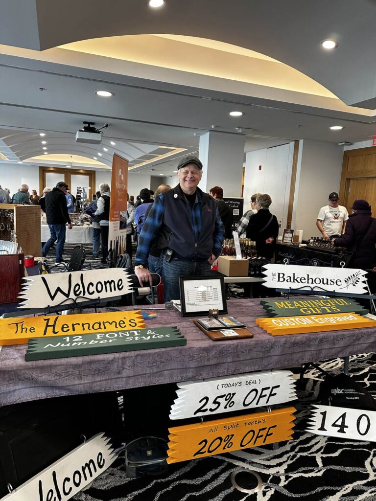 Smiling vendor with hand-painted wooden signs at the indoor Osoyoos Farmers’ Market